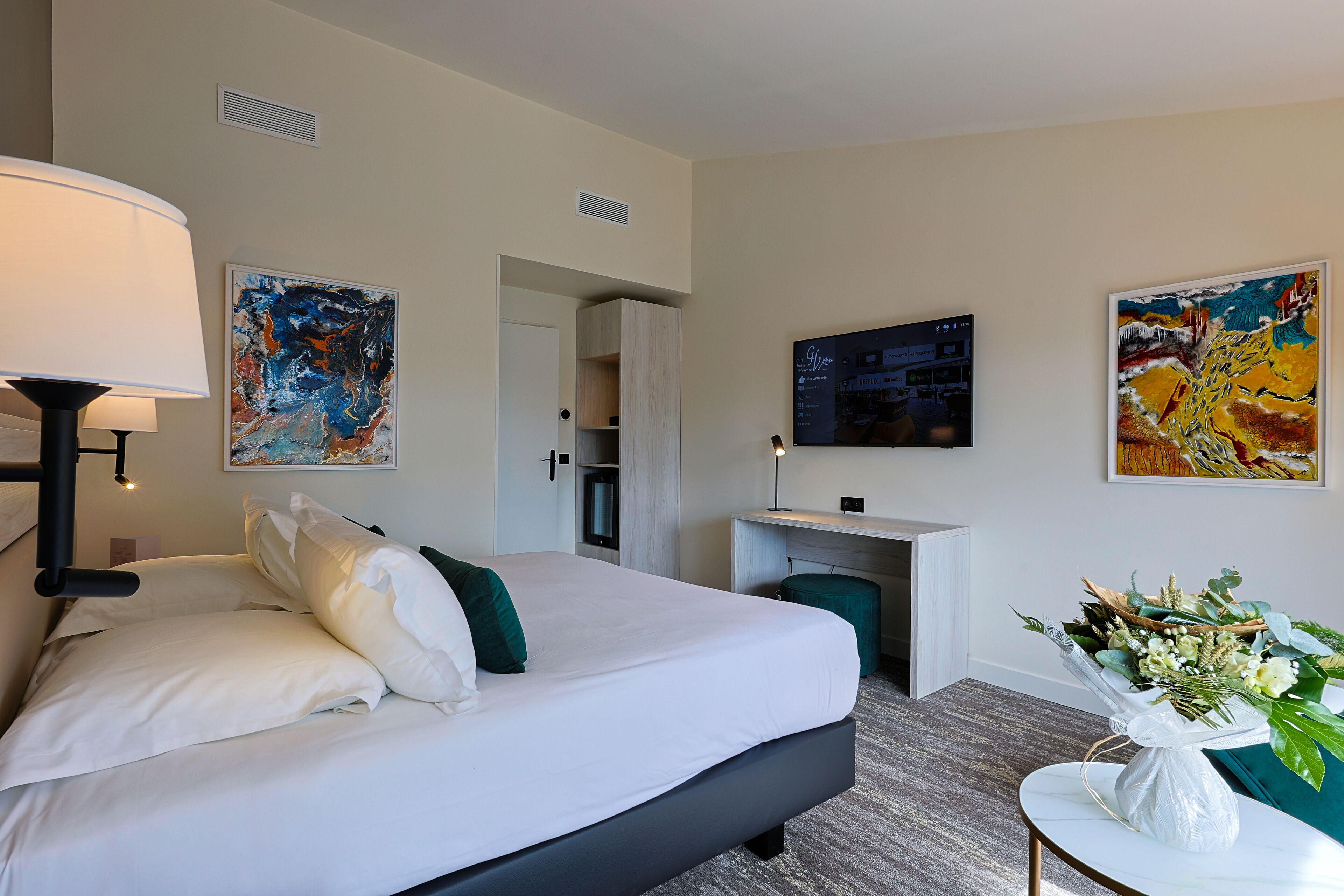 A hotel room with a bed, a desk, a TV, and artwork on the walls.