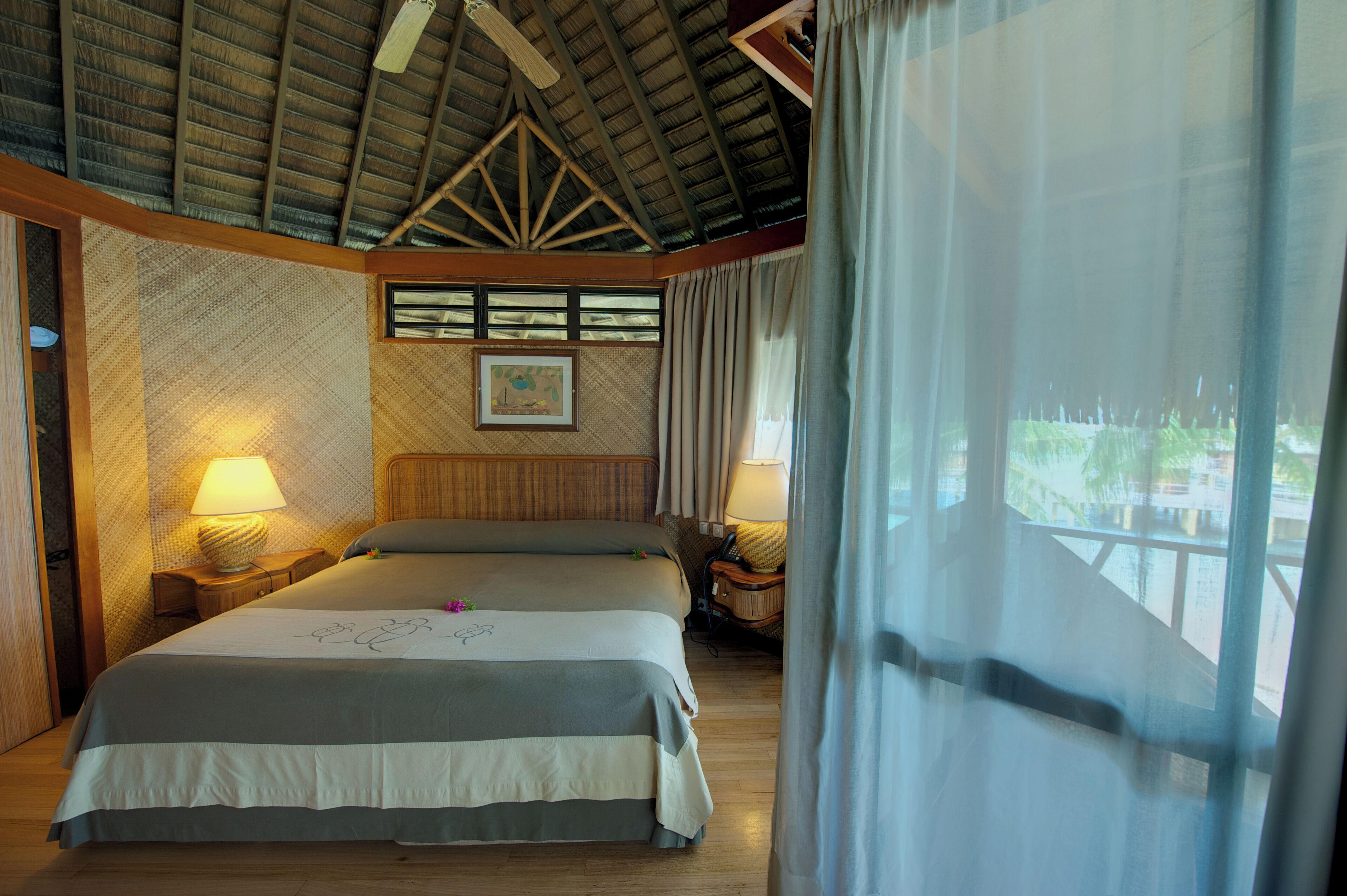 A spacious bedroom with a large bed, wooden headboard, and a ceiling fan. There are two bedside tables with lamps and a window with curtains.