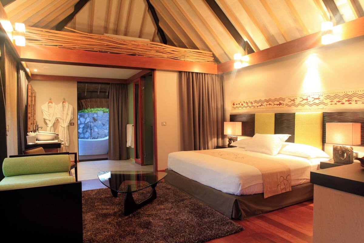 Quarto de hotel com uma cama, área de estar com um sofá verde, mesa de centro de vidro e banheiro com pia e espelho.