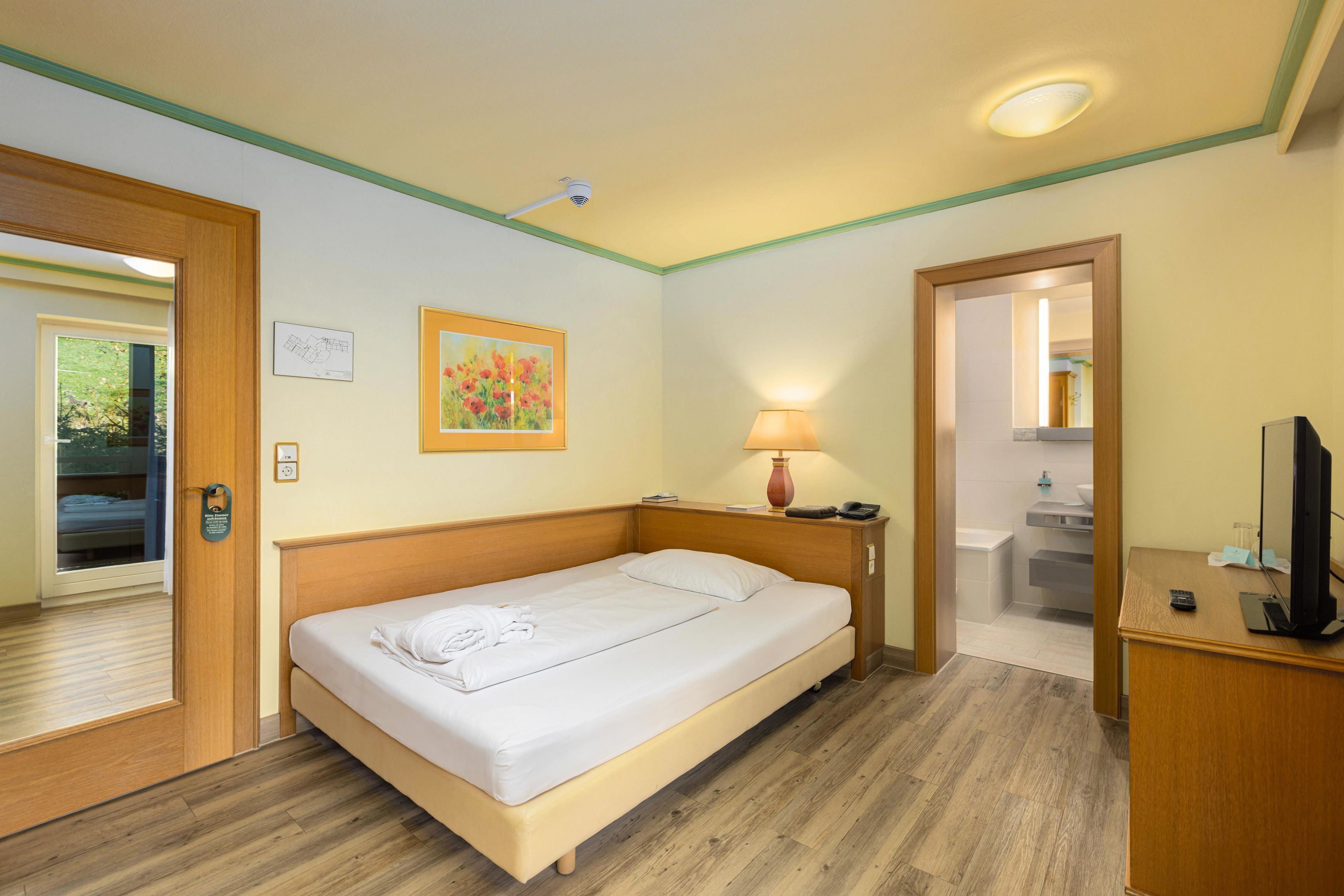 A single bed with a wooden headboard, a bedside lamp, a desk with a computer, and a bathroom visible through an open door.