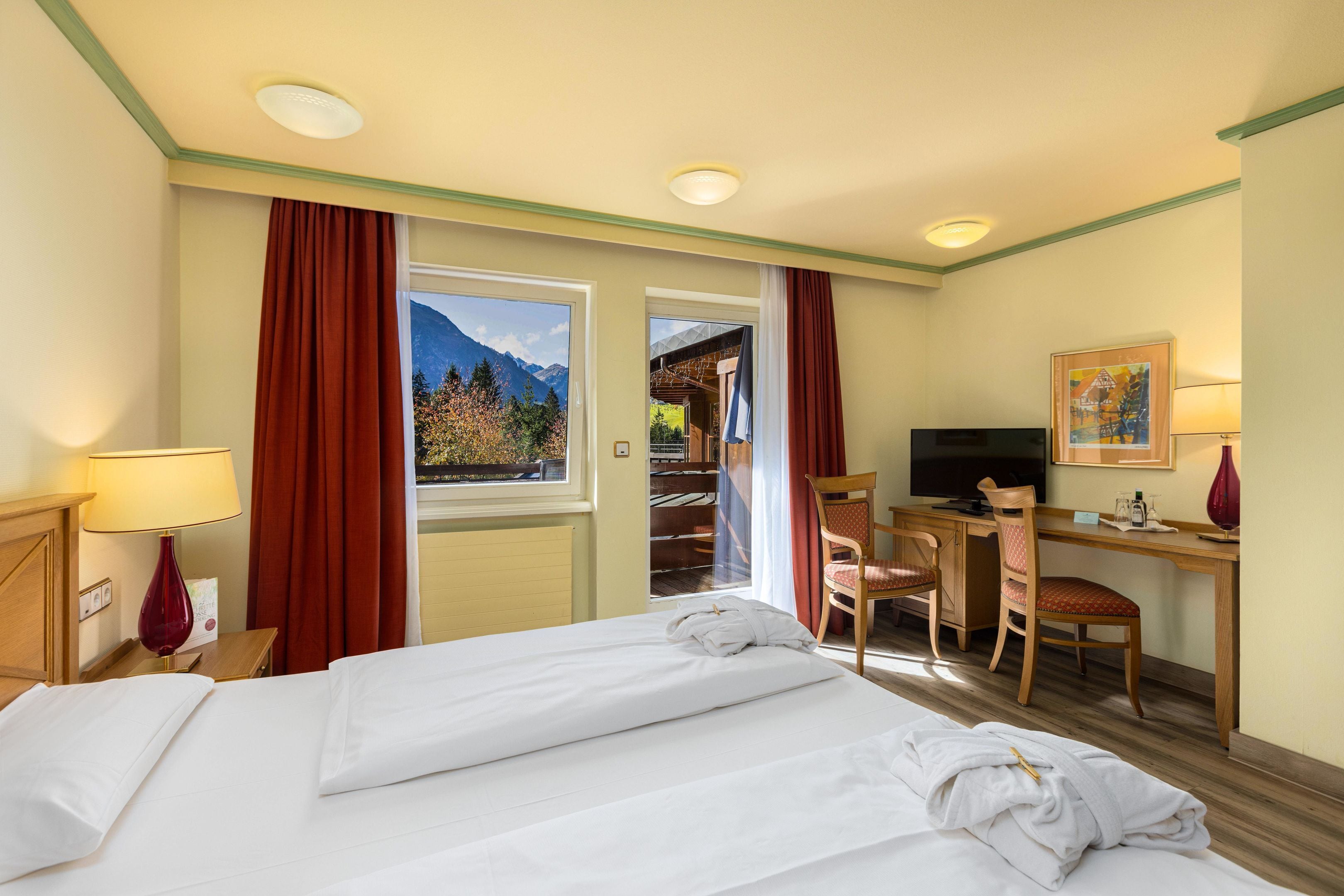 A hotel room with a bed, desk, chair, TV, and a view of mountains.