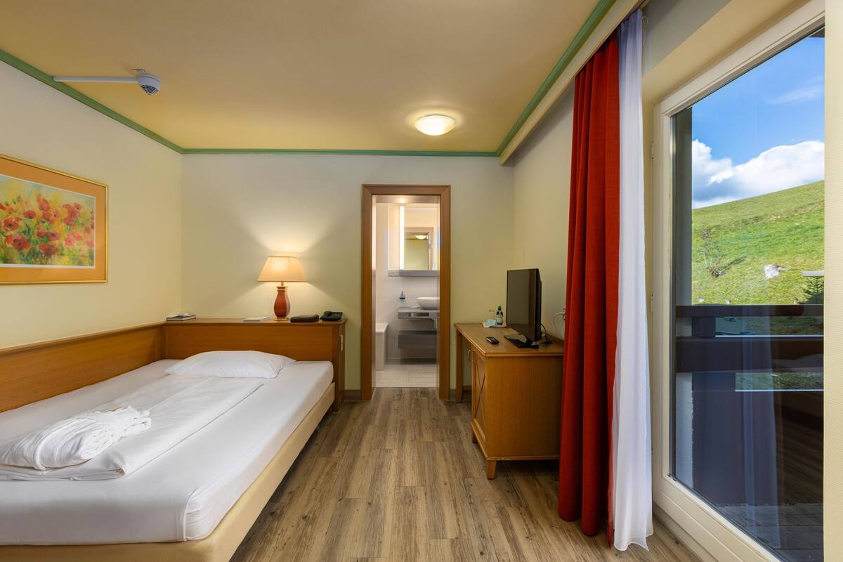 A hotel room with a bed, a desk with a TV, a window with curtains, a painting on the wall, and a hallway leading to a bathroom.