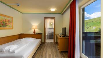 Habitación de hotel con cama, escritorio con televisor, ventana con cortinas, un cuadro en la pared y un pasillo que da a un baño.