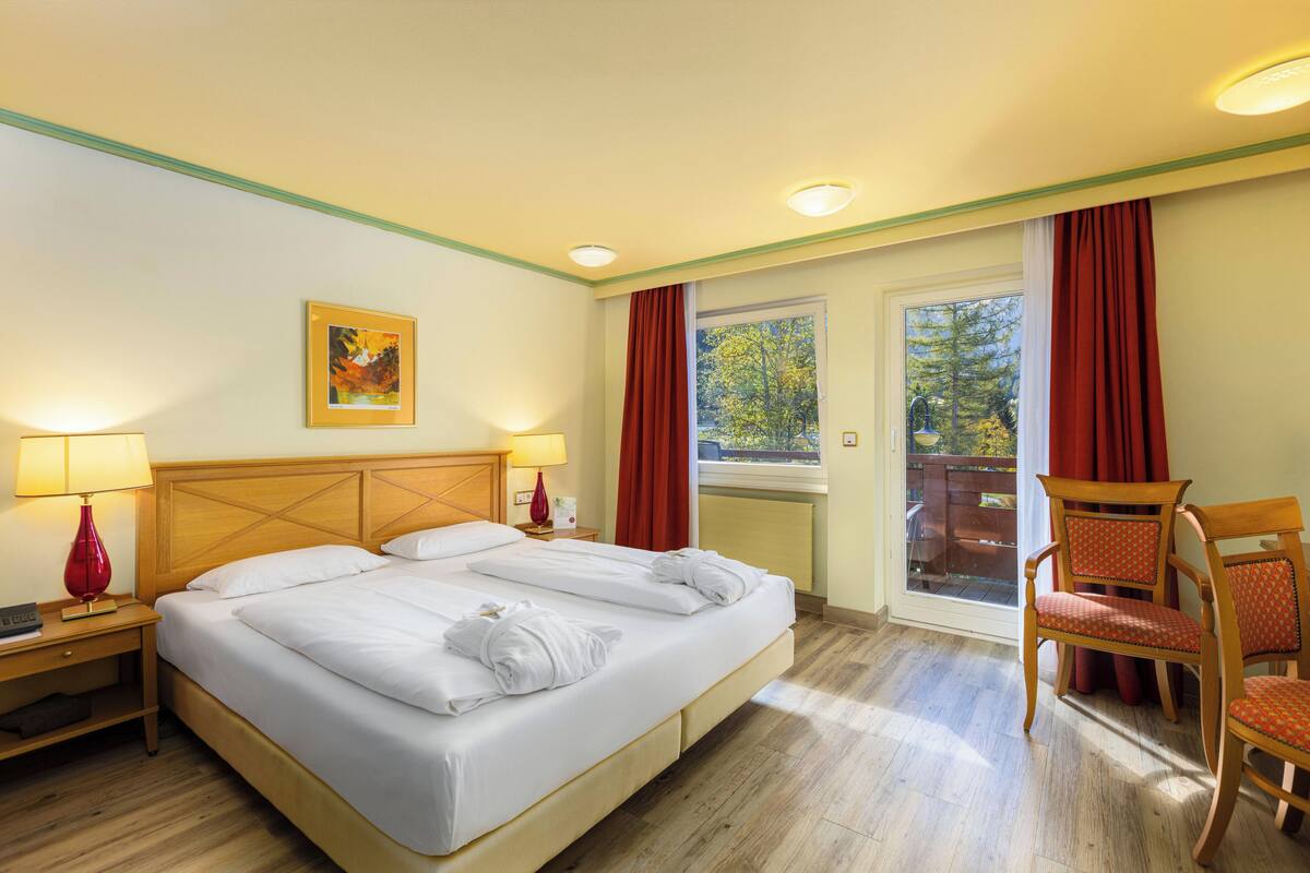 A hotel room with a double bed, two bedside tables with lamps, a window with curtains, a chair, and a painting on the wall.