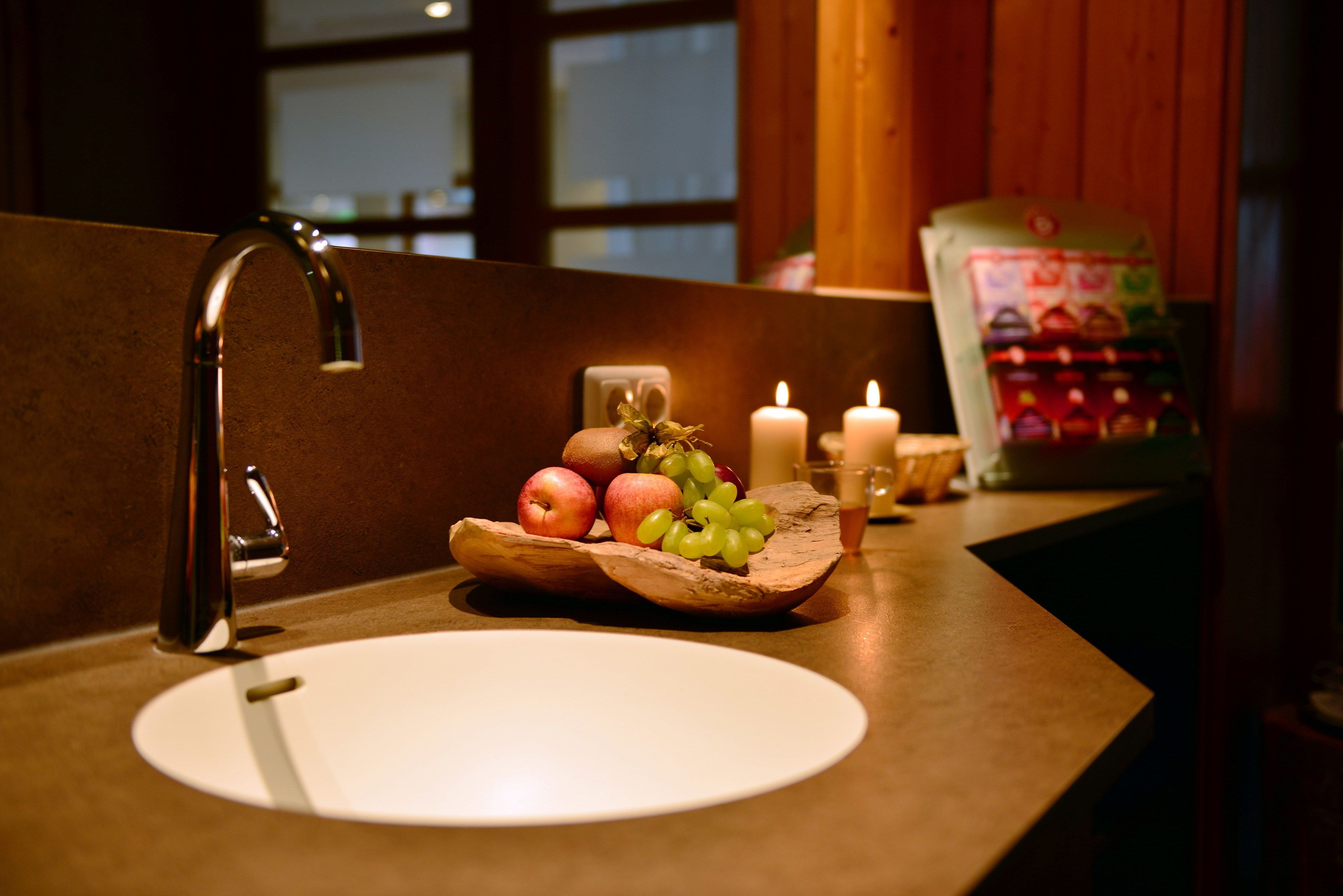 Un lavabo de salle de bain avec un récipient en bois contenant des pommes et des raisins, une bougie et un paquet de produits de toilette.
