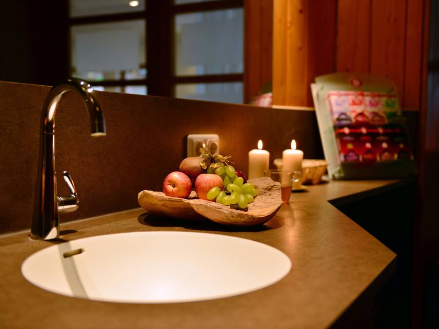 Un lavabo de salle de bain avec un récipient en bois contenant des pommes et des raisins, une bougie et un paquet de produits de toilette.