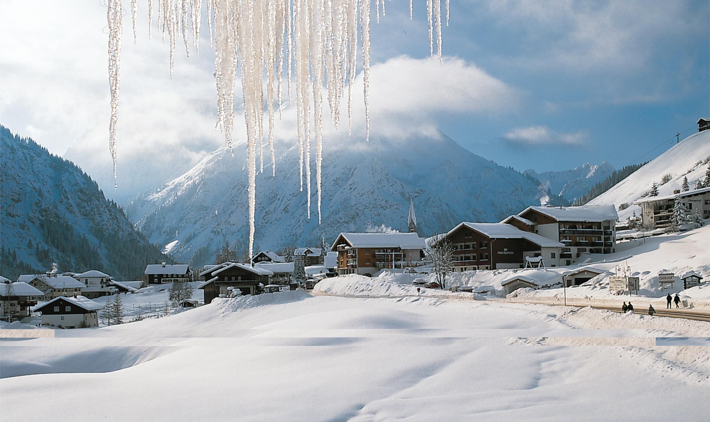 Un village de montagne enneigé, avec des bâtiments en bois et une église, entouré de collines recouvertes de neige.