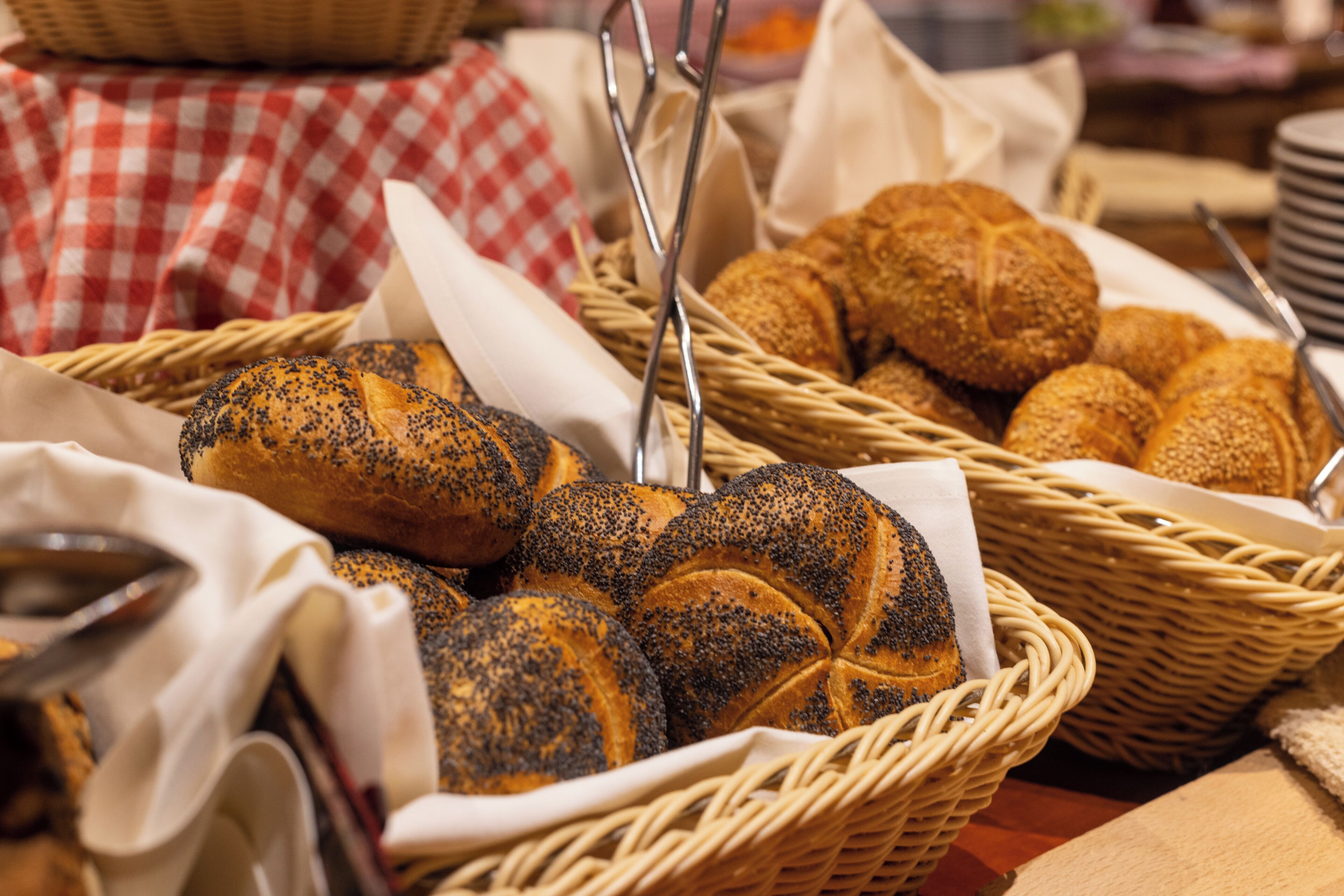 A basket of bread rolls with poppy seeds, a checkered cloth, and a metal spoon.