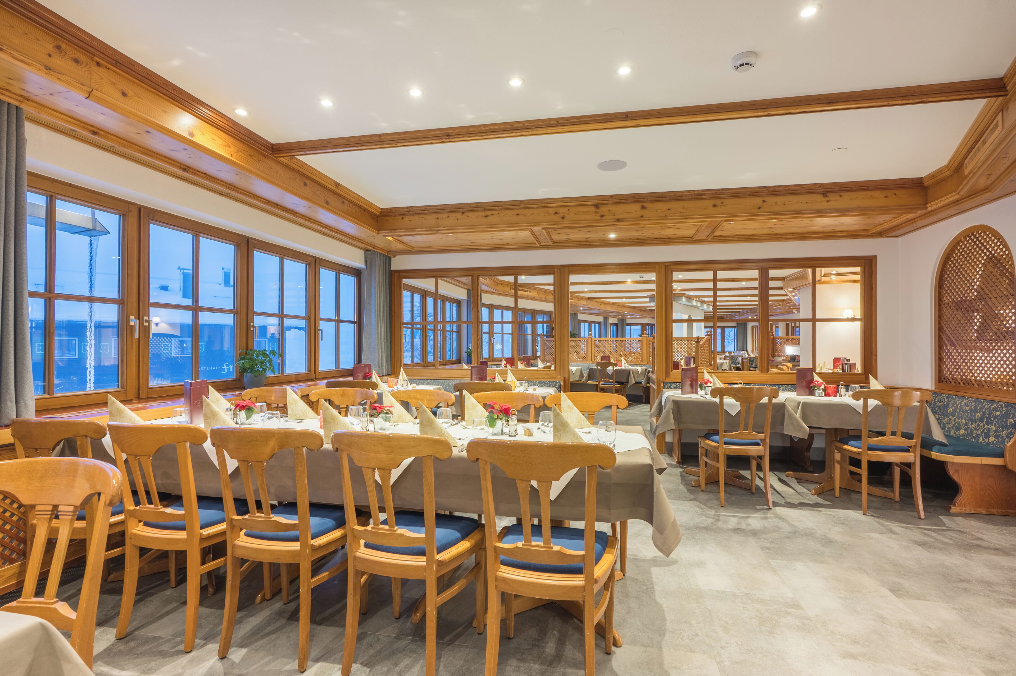 A spacious dining area with wooden tables and chairs, large windows, and a high ceiling with wooden beams.