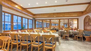 A spacious dining area with wooden tables and chairs, large windows, and a high ceiling with wooden beams.