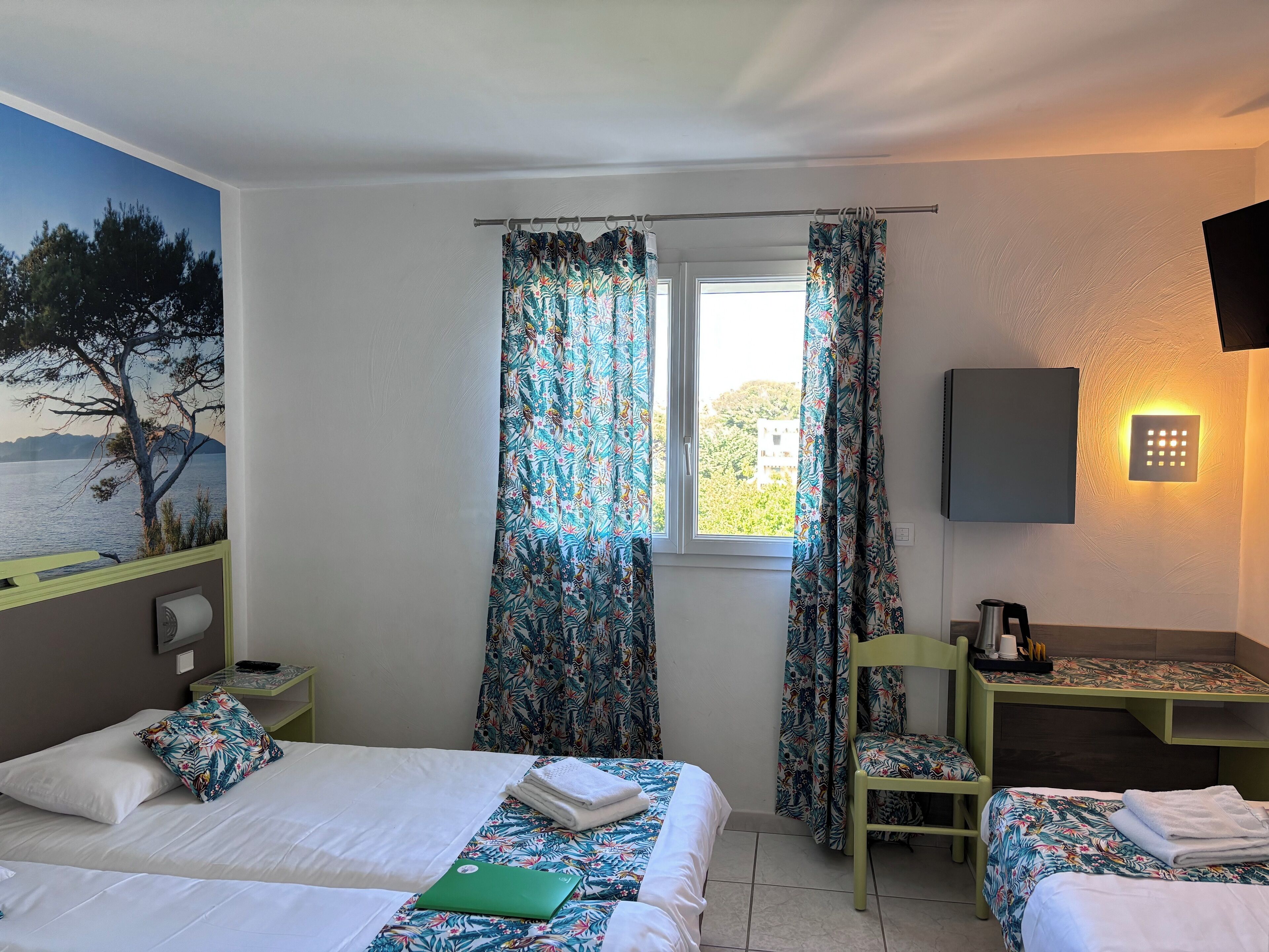 A hotel room with two beds, a desk, a chair, a view of the sea, and a wall-mounted light.