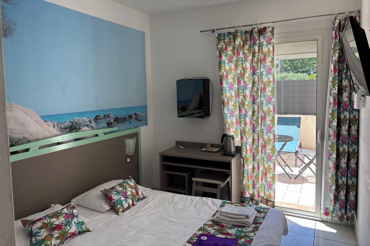 A bedroom with a bed, a desk, a chair, a painting of a beach, and a view of a pool through a window.