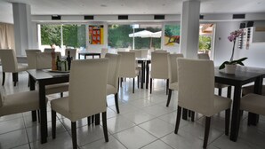 A modern dining area with multiple tables and chairs, large windows with views of greenery, and artwork on the walls. - Hôtel de la Mer (Vallauris)