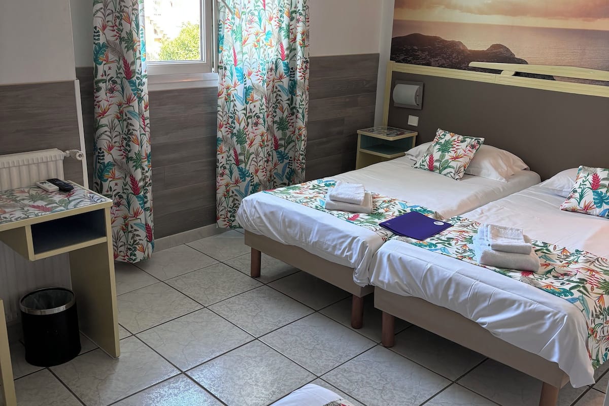 A hotel room with two beds, a desk, a waste bin, and a window with curtains.