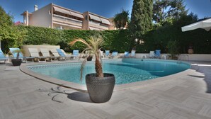 A swimming pool with lounge chairs, a palm tree planter, and a building in the background. - Hôtel de la Mer (Vallauris)