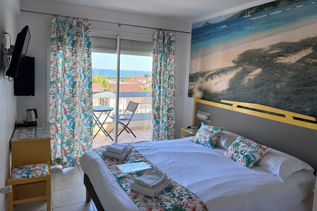 A hotel room with a large bed, a small table with a kettle, a chair, and a balcony with a view of the sea.