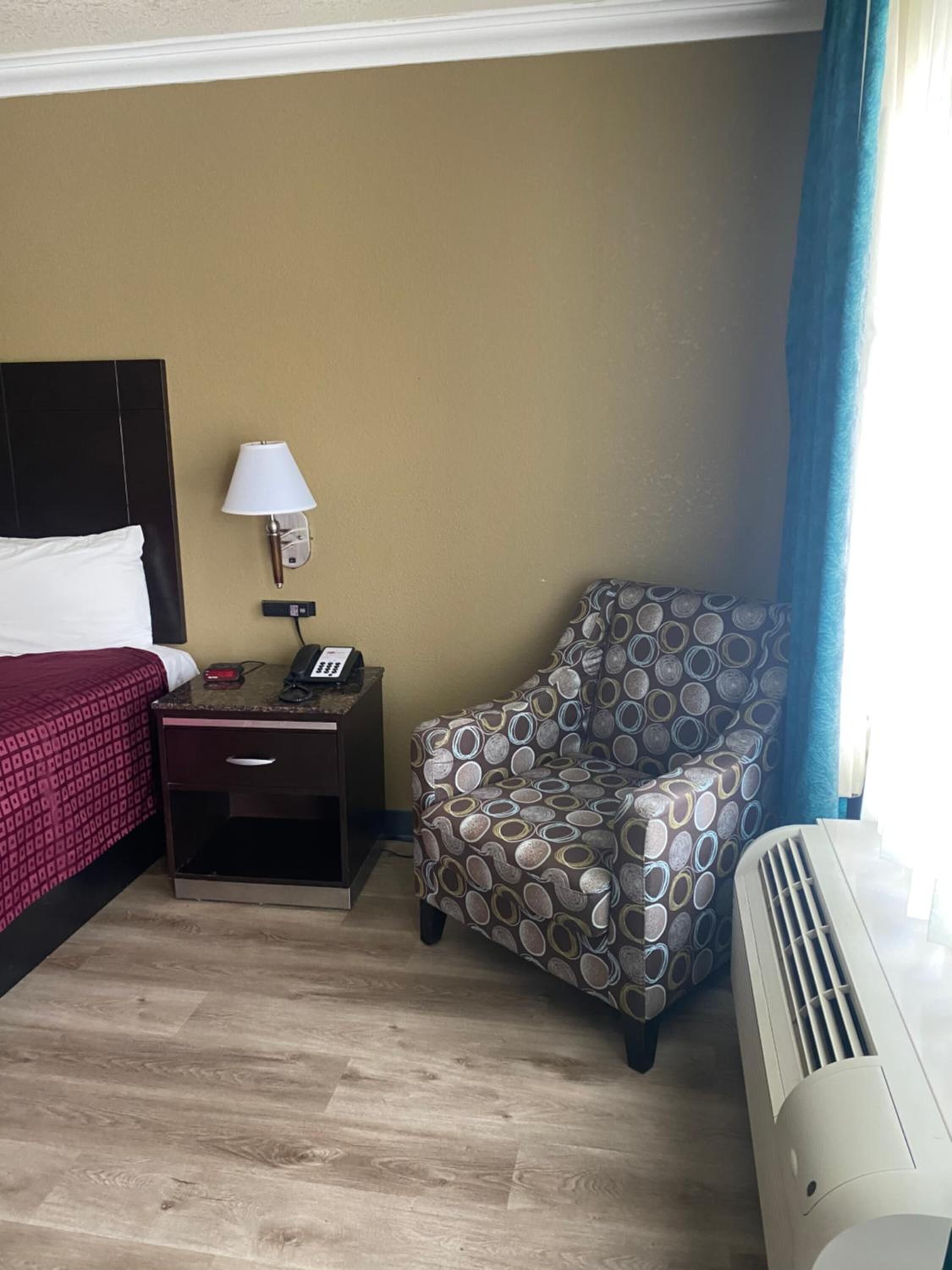 A hotel room with a bed, bedside table, lamp, chair, and air conditioning unit.