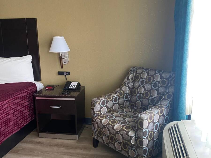 A hotel room with a bed, bedside table, lamp, chair, and air conditioning unit.