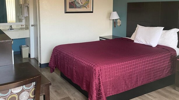 A hotel room with a bed, a desk, a chair, a nightstand, a lamp, and a painting on the wall. - Atascadero Inn (Atascadero)