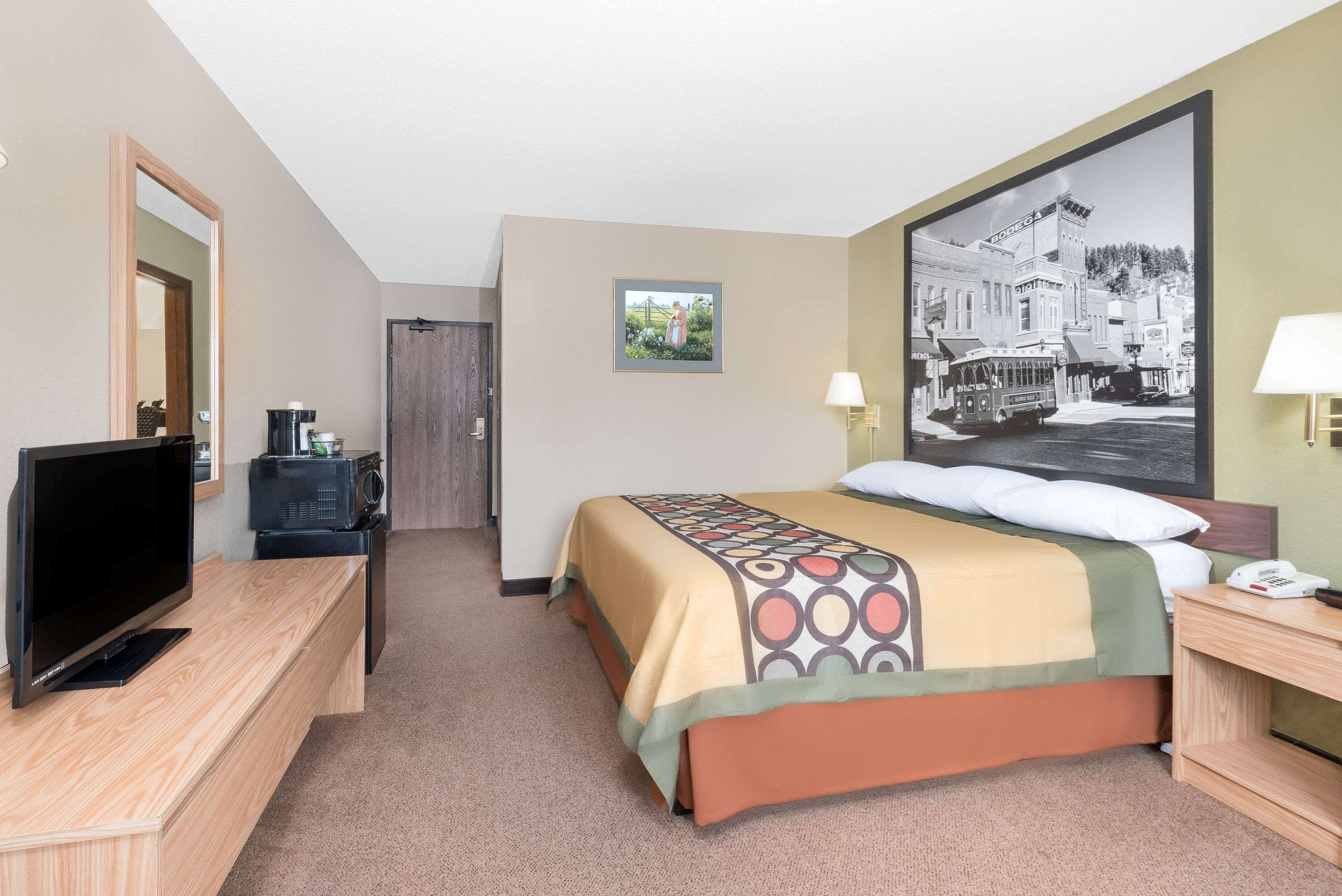 Habitación de hotel con cama, televisor, escritorio, lámpara y un cuadro en la pared.