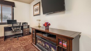 A hotel room with a desk, chair, flat-screen TV, framed picture, and a fireplace with books. - Colstrip Inn & Suites (Colstrip)