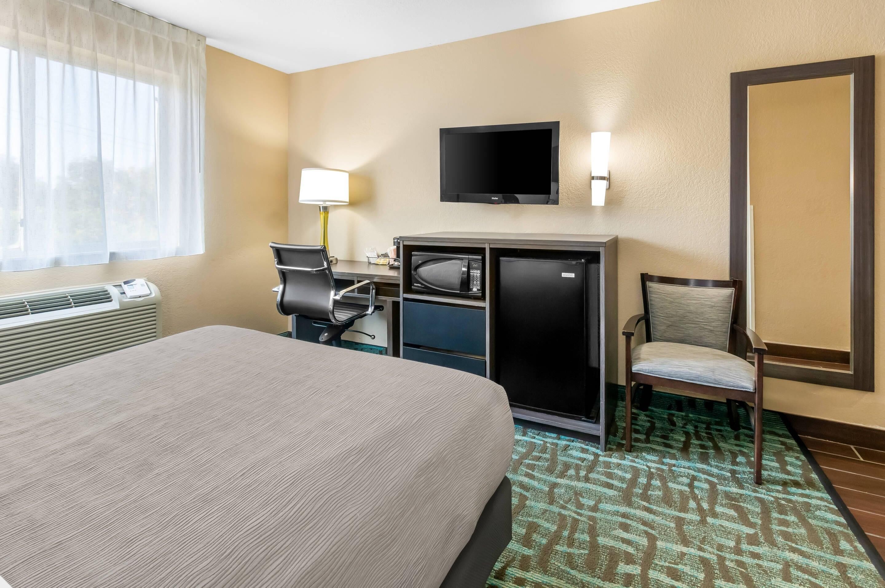 A hotel room with a bed, a desk with a computer and a microwave, a TV, and a chair.