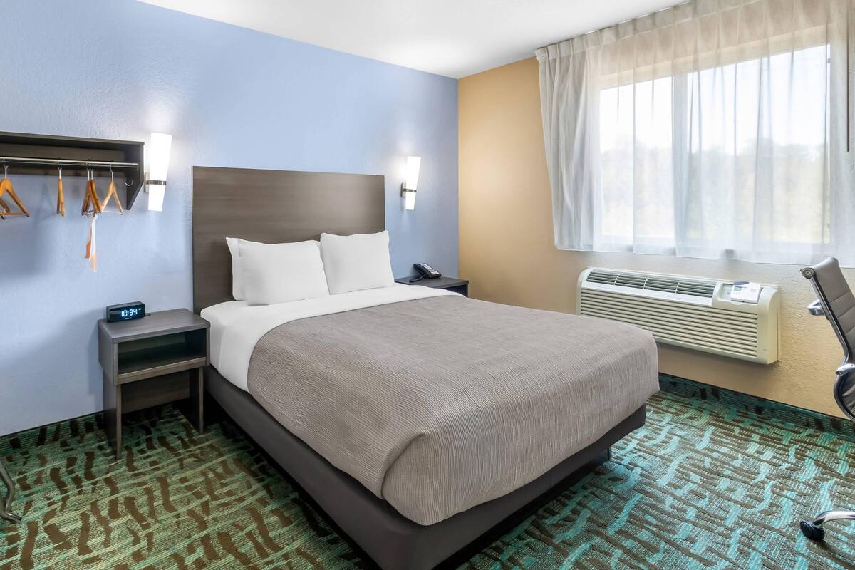 Une chambre d’hôtel comprenant un lit, une table de chevet, une lampe fixée au mur, une fenêtre avec des rideaux et un climatiseur fixé au mur.