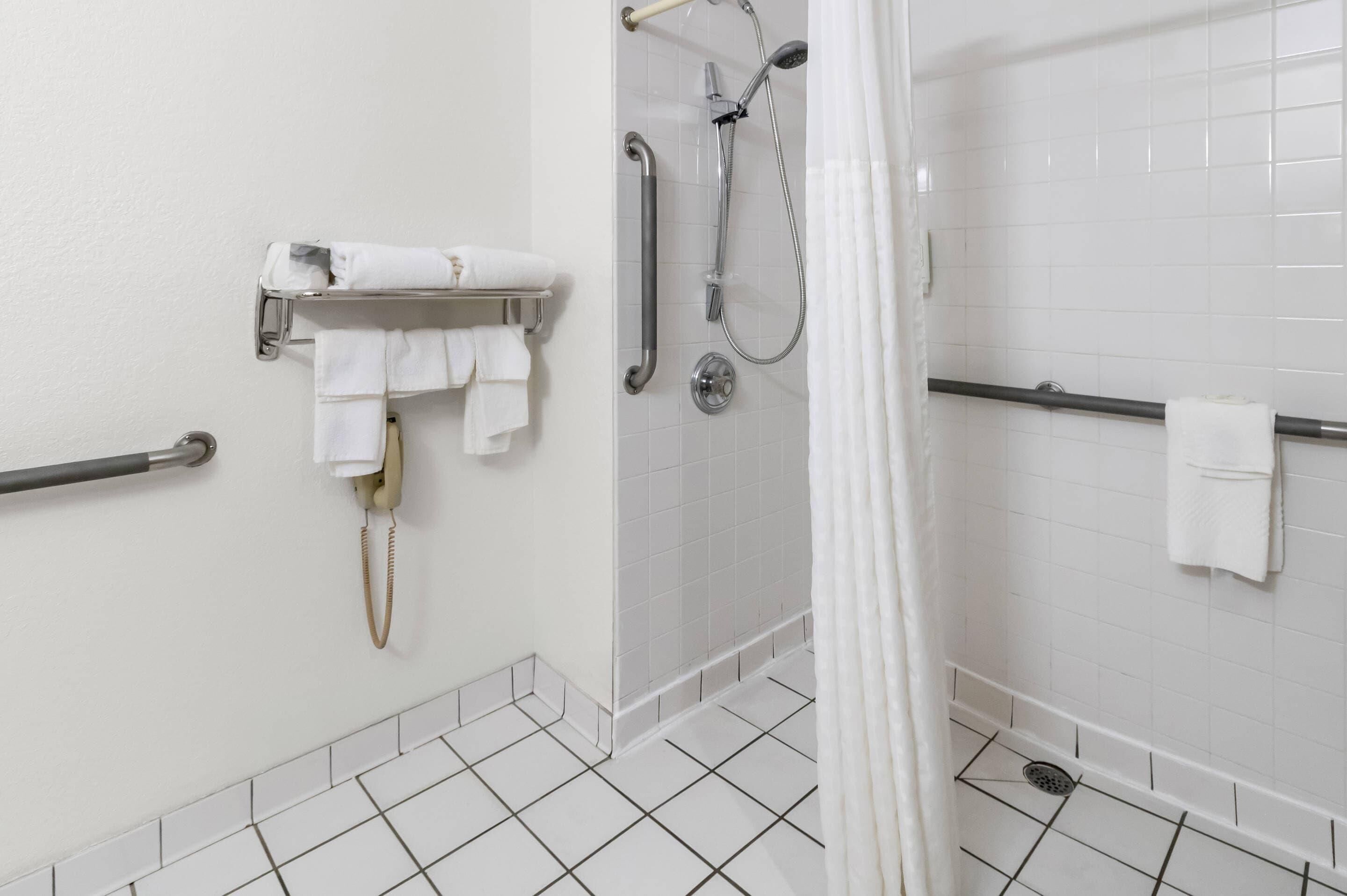 Ein Badezimmer mit weißen Fliesenwänden, einer Dusche mit Duschhandstück, einer Haltestange und einem Handtuchhalter mit mehreren Handtüchern.