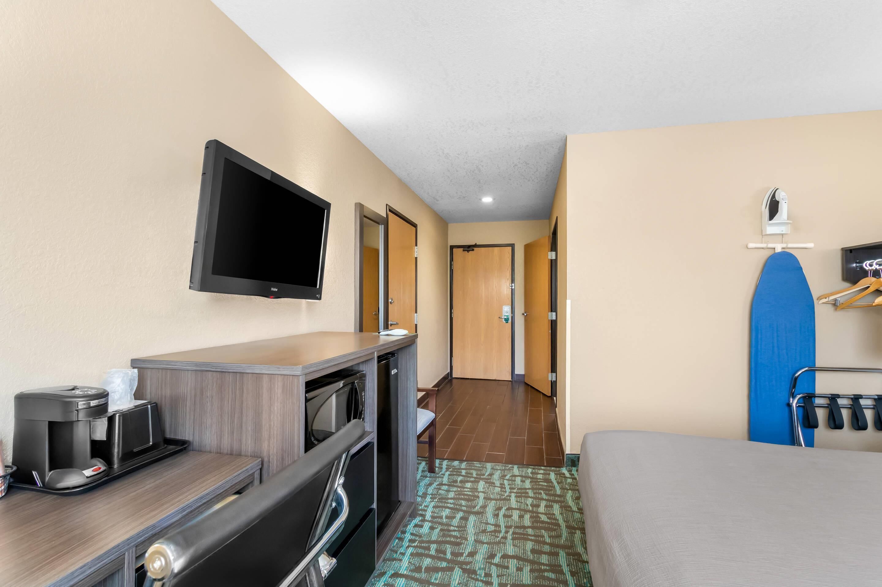 A hotel room with a desk, a flat-screen TV, a coffee maker, a microwave, a mini-fridge, a bed, a shower, and a blue surfboard on a rack.
