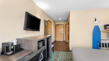 A hotel room with a desk, a flat-screen TV, a coffee maker, a microwave, a mini-fridge, a bed, a shower, and a blue surfboard on a rack.