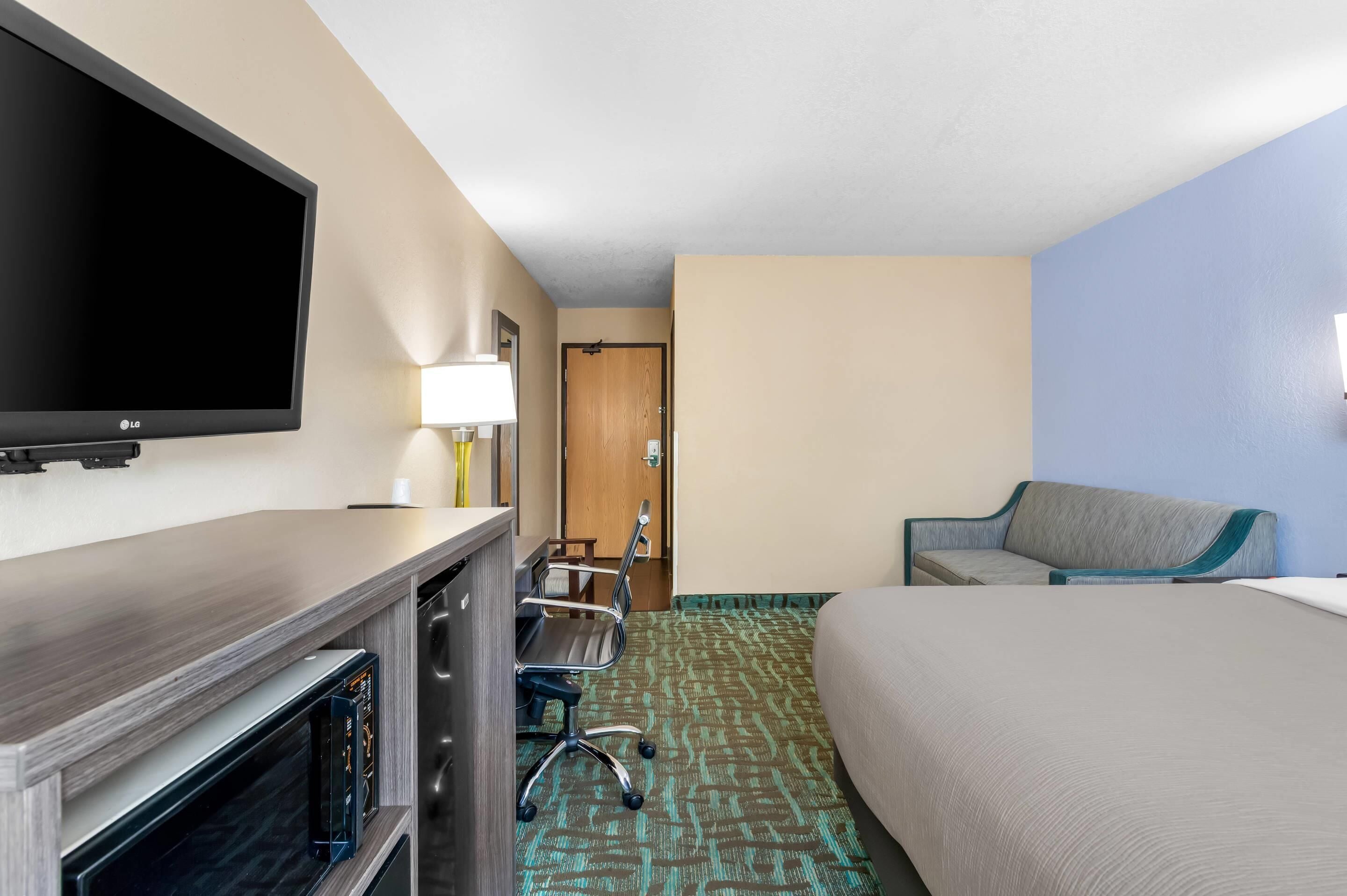 A hotel room with a flat-screen TV, a desk with a lamp, a chair, a bed, and a door leading to a bathroom.