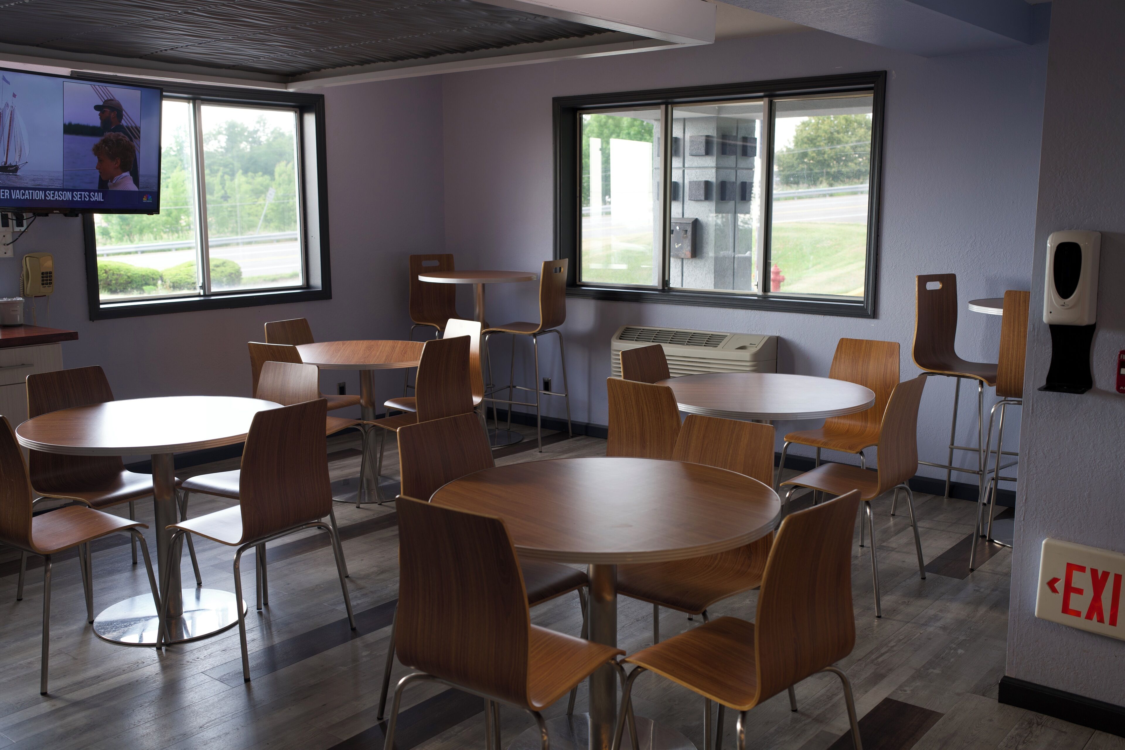 Un salón de conferencias con varias mesas y sillas, ventanas amplias y una señal de salida.