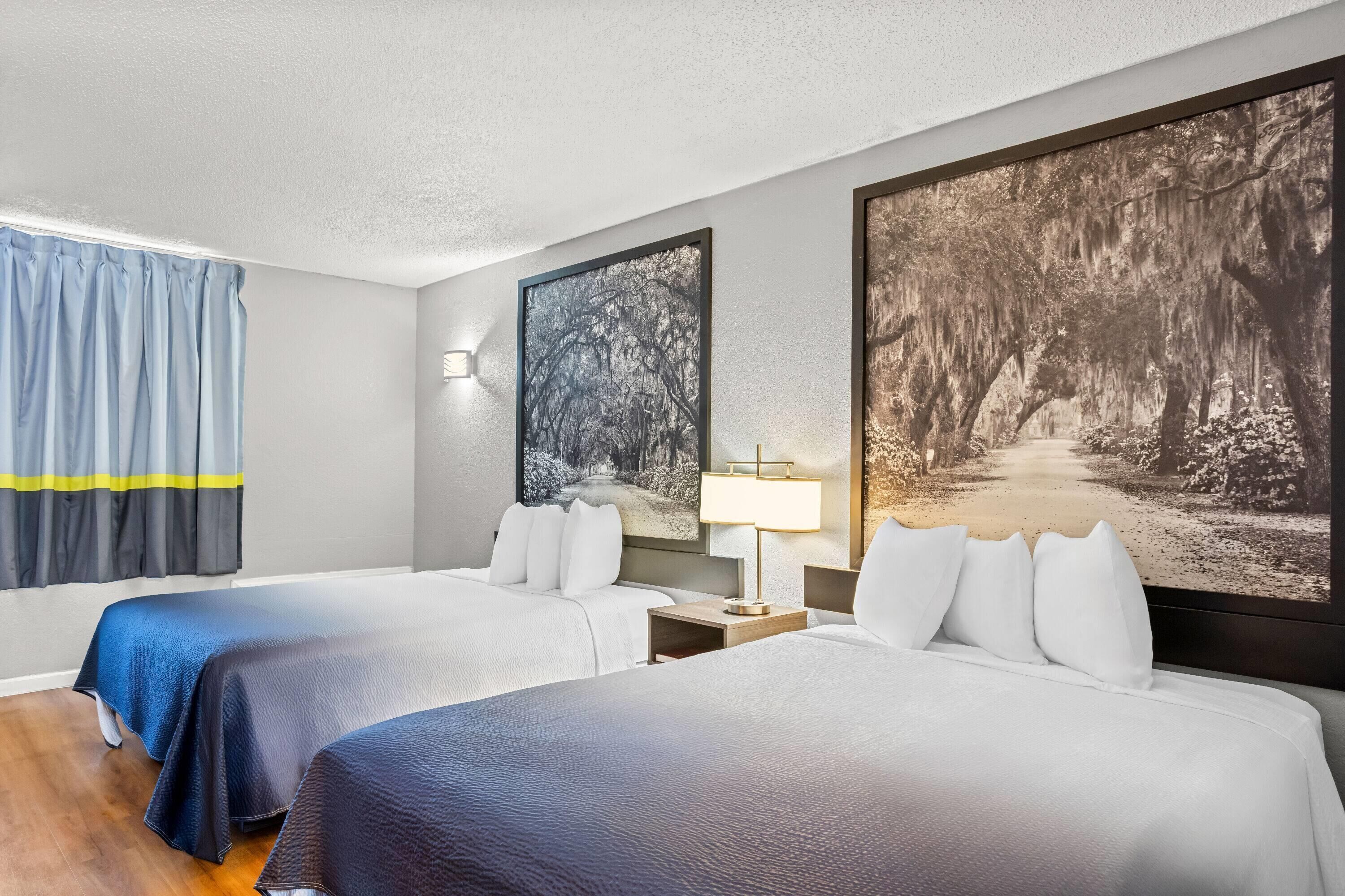 Habitación de hotel con dos camas, una mesita de noche, una lámpara y dos grandes cuadros con motivos forestales en las paredes.