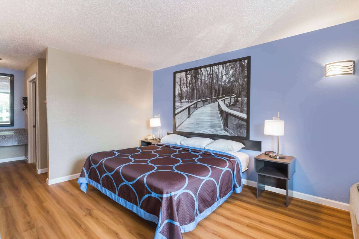 Ein Schlafzimmer mit einem großen Bett, zwei Nachttischen, einem Beistelltisch, einem Bild an der Wand und einer Tür zu einem anderen Raum.