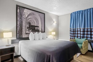 Habitación de hotel con una cama grande, dos lámparas de noche, una lámpara de pared, cortina estampada y un cuadro enmarcado sobre la cama.