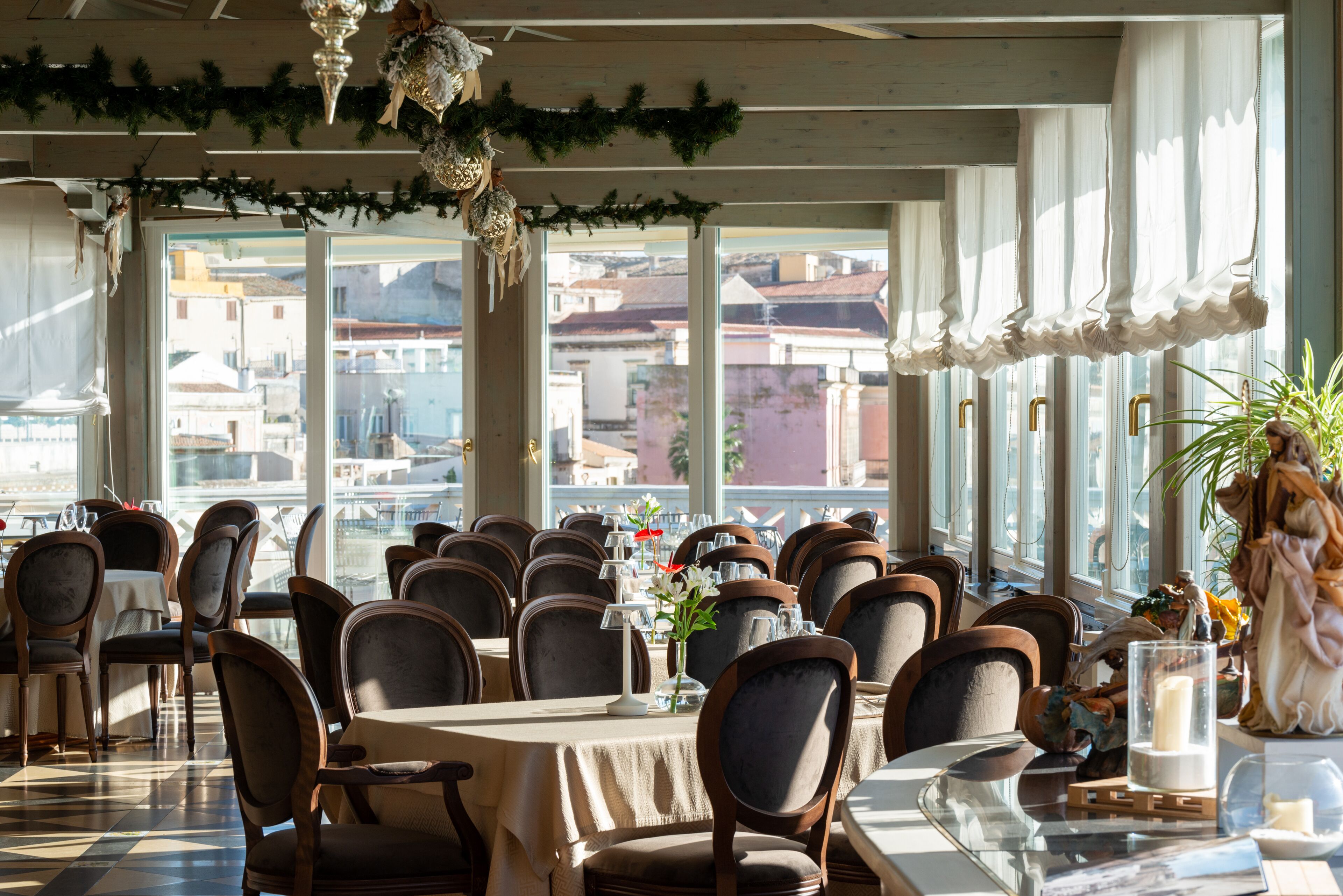 Un restaurant doté de nombreuses tables et chaises, de grandes fenêtres offrant une vue sur les bâtiments, et d’éléments décoratifs tels que des guirlandes et une statue.
