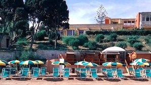 Un área junto a la piscina con sillones de descanso y sombrillas, una piscina cristalina y un edificio con múltiples ventanas al fondo.