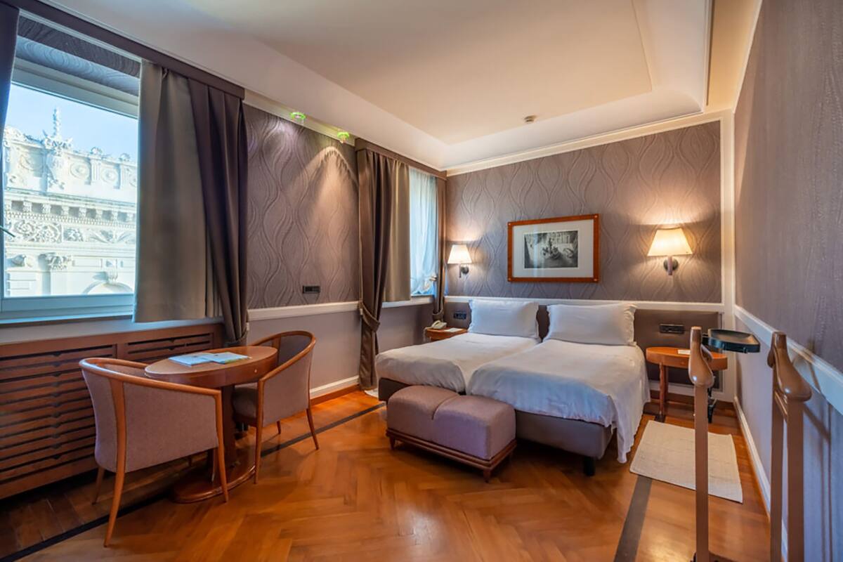 Una habitación de hotel con cama, escritorio, silla, mesita de noche y ventana con cortinas.