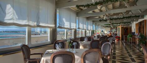 A restaurant with tables and chairs, large windows, and a view of the sea.
