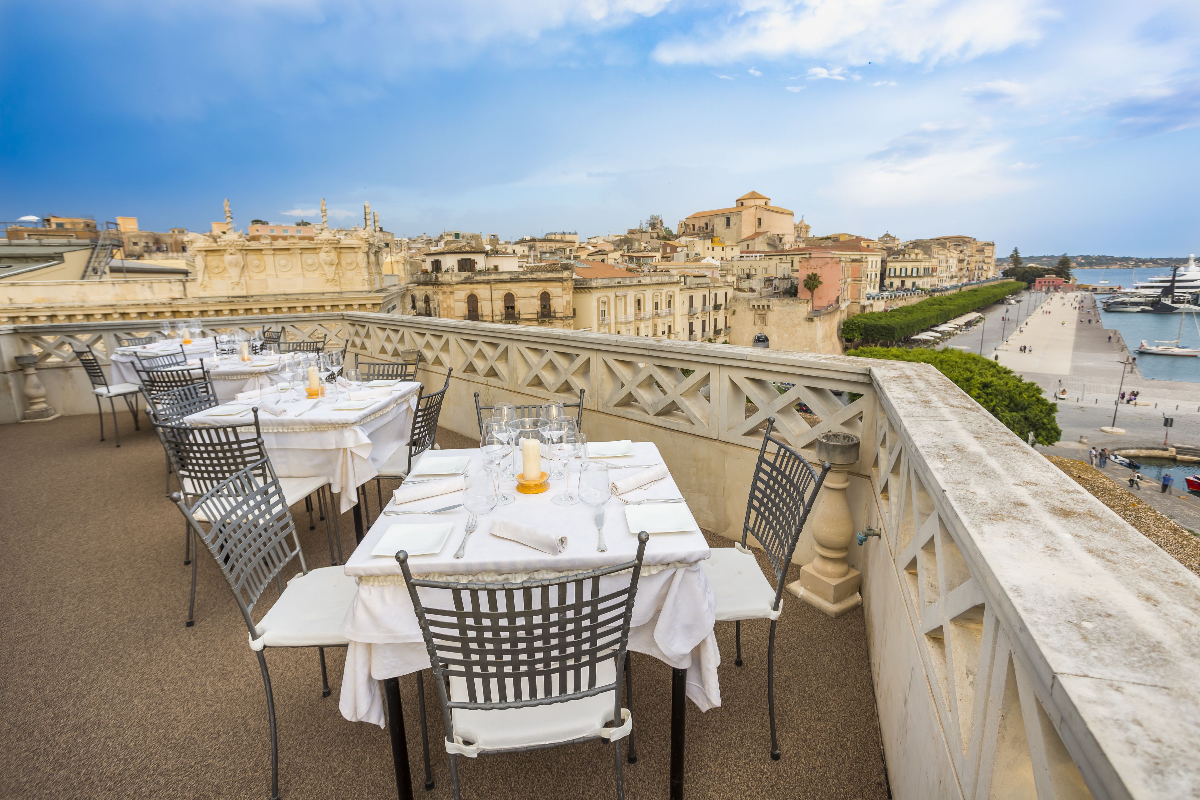 Une terrasse sur le toit, avec des tables dressées pour les repas, offrant une vue imprenable sur un paysage urbain historique et la mer.