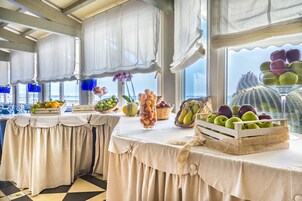 A dining area with tables set for service, featuring a variety of fruits and a view of the sea through large windows.
