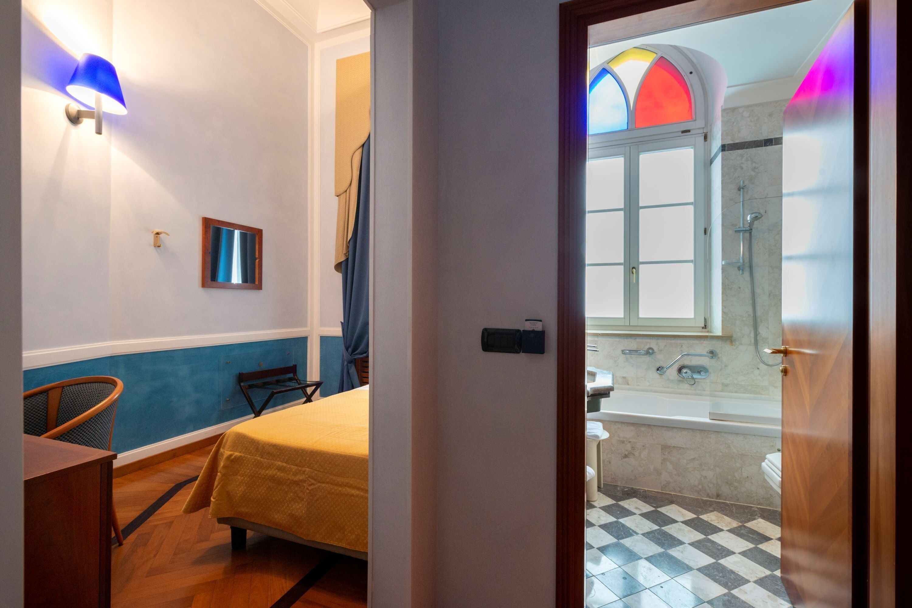 A hotel room with a bed, a chair, a small table, a window with stained glass, and a bathroom with a bathtub and shower.