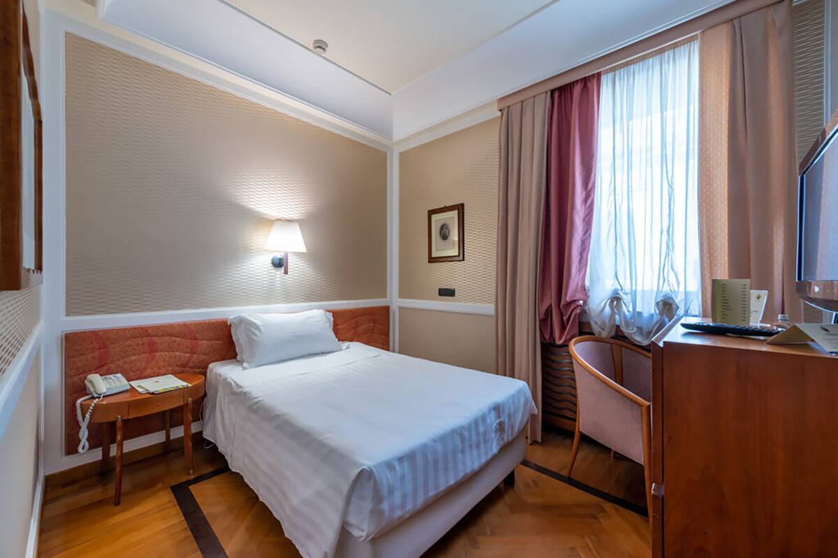 Habitación de hotel con cama, mesita de noche con teléfono, escritorio con silla, lámpara, cuadro en la pared y ventana con cortinas.