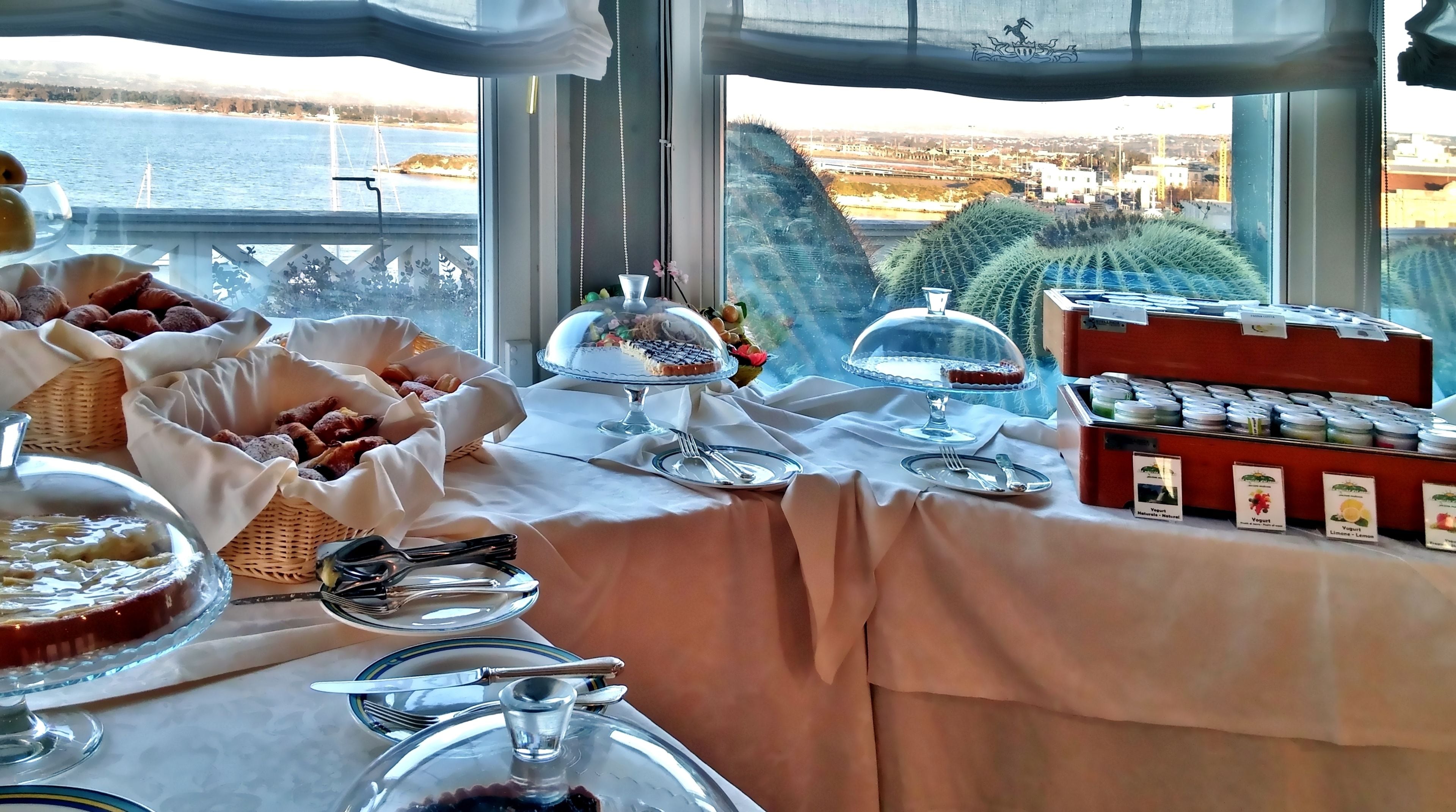 Un bufé de desayuno con diversas opciones de comida, vista al mar y al paisaje urbano a través de amplias ventanas, y una mesa dispuesta con cubiertos y platos.