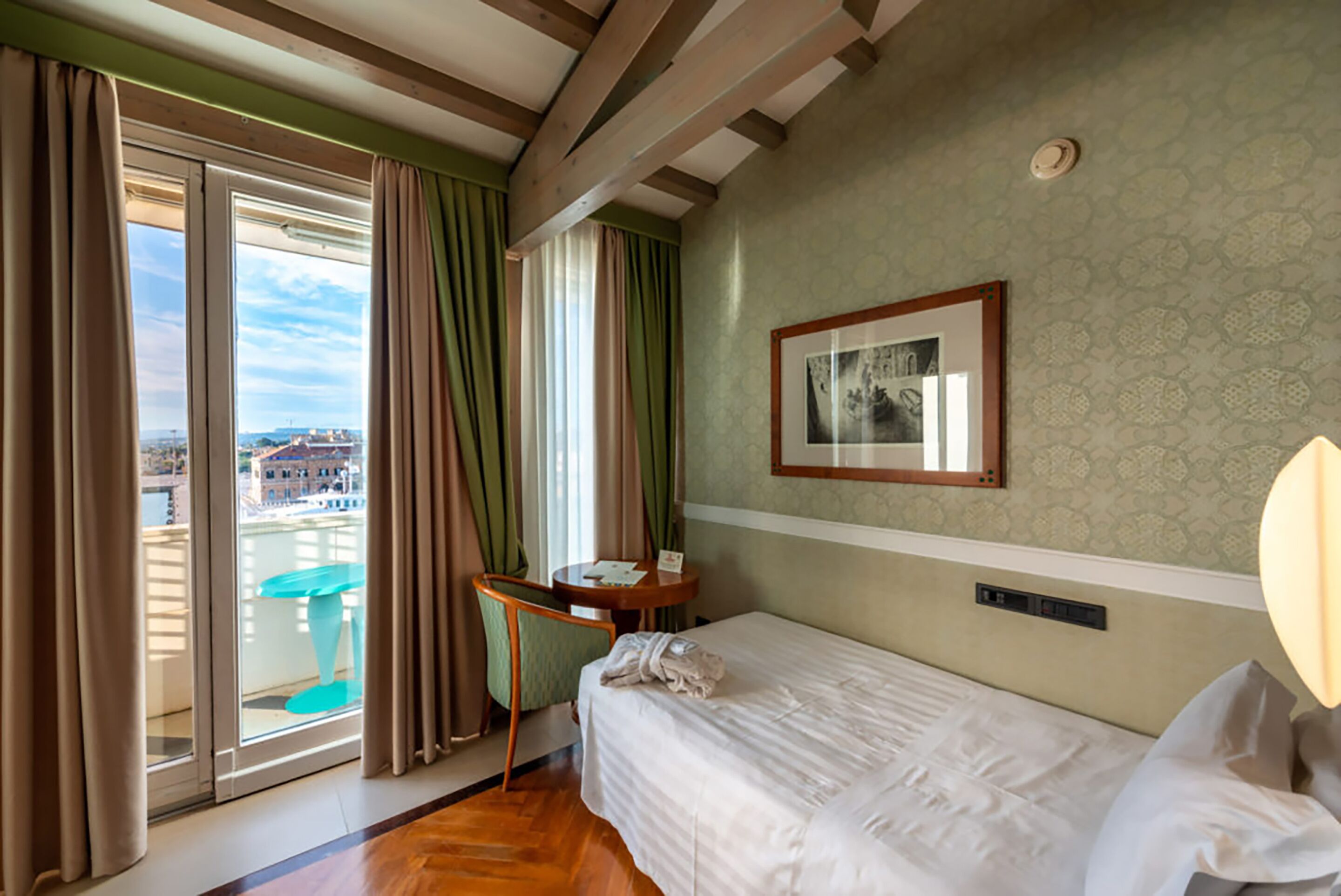 A hotel room with a bed, a small table, a chair, a window with curtains, and a framed picture on the wall.