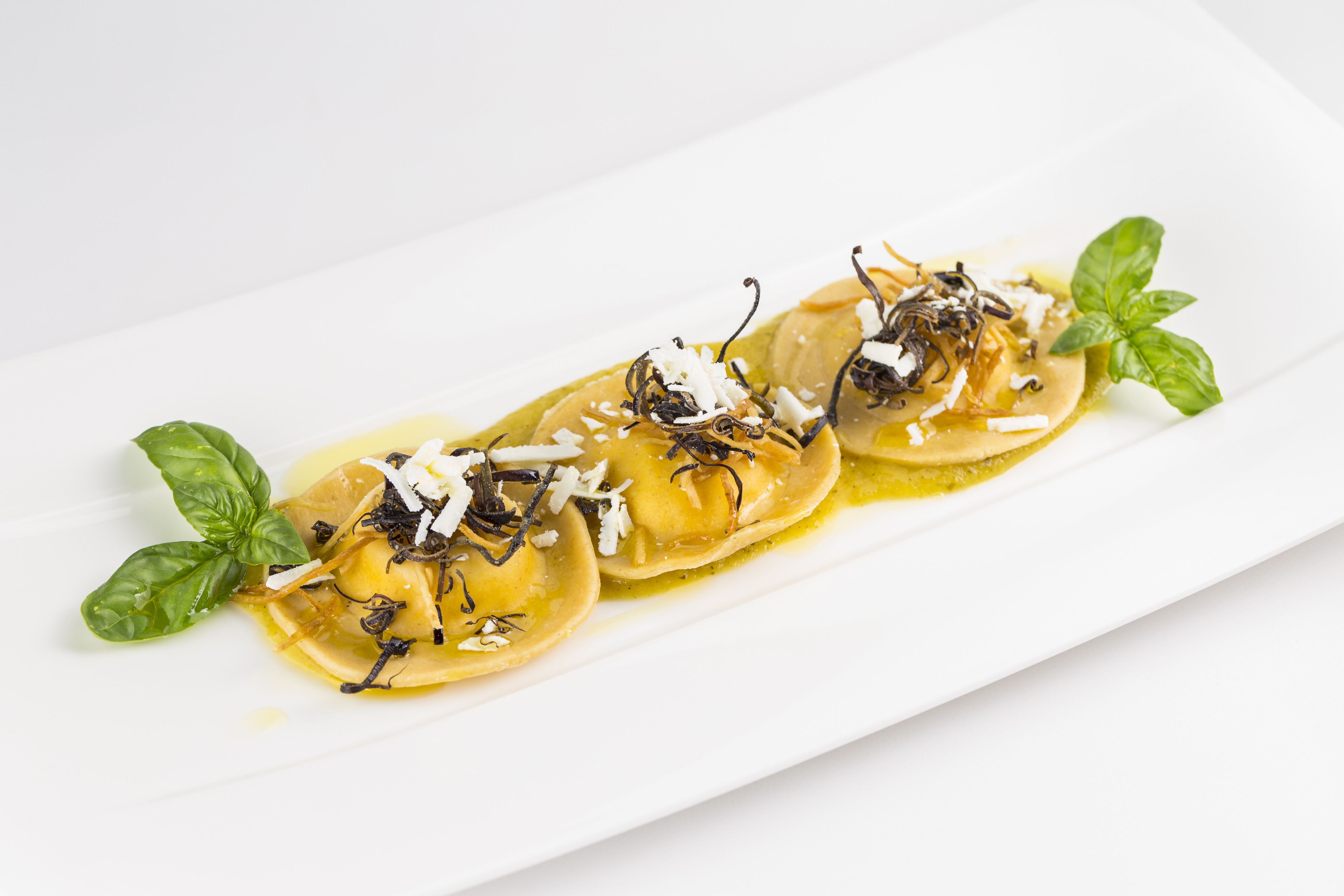 Un plato rectangular blanco con tres ravioles adornados con hierbas y queso, acompañado de hojas de albahaca.