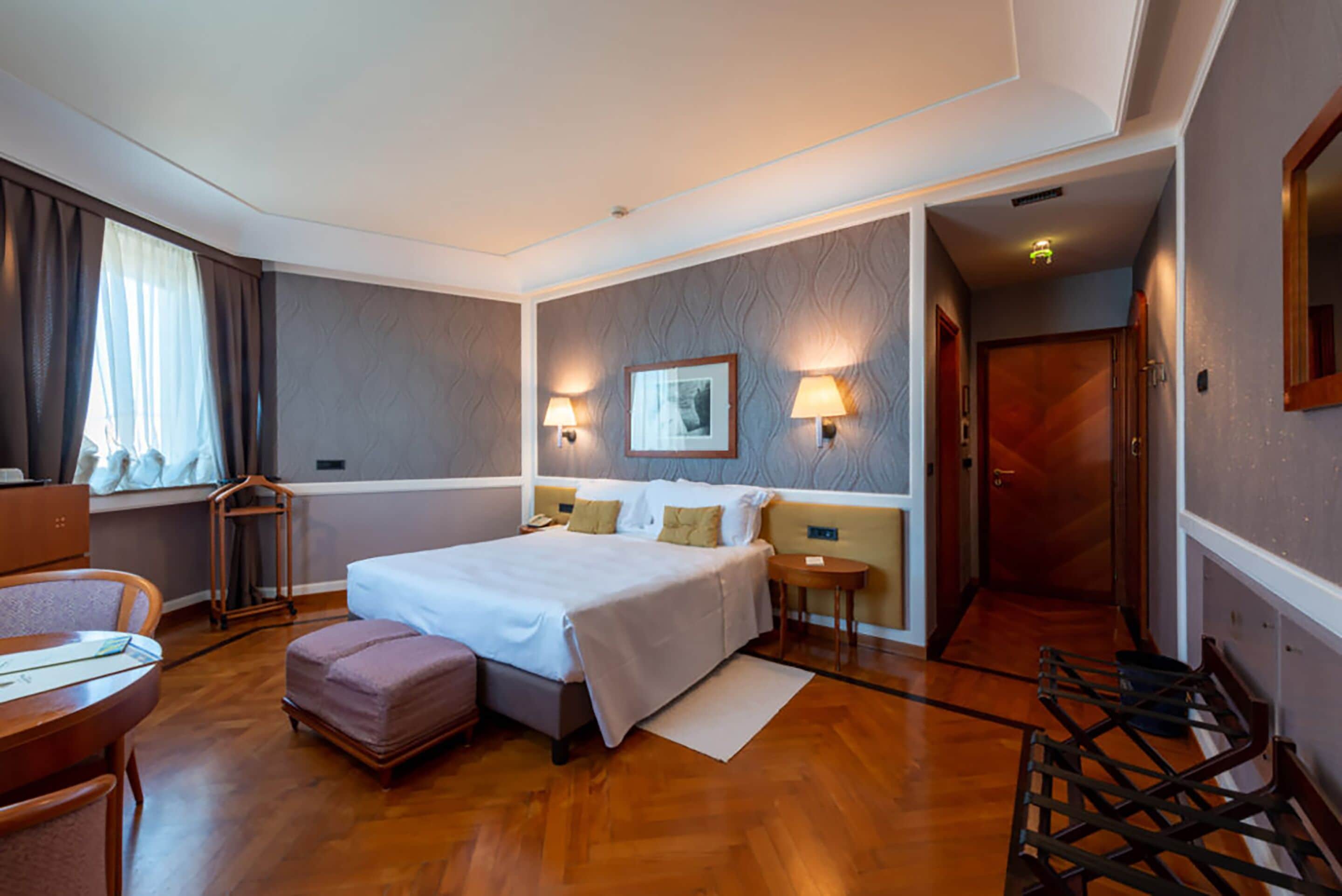 A hotel room with a large bed, bedside tables, a desk, a chair, a window with curtains, and a door.