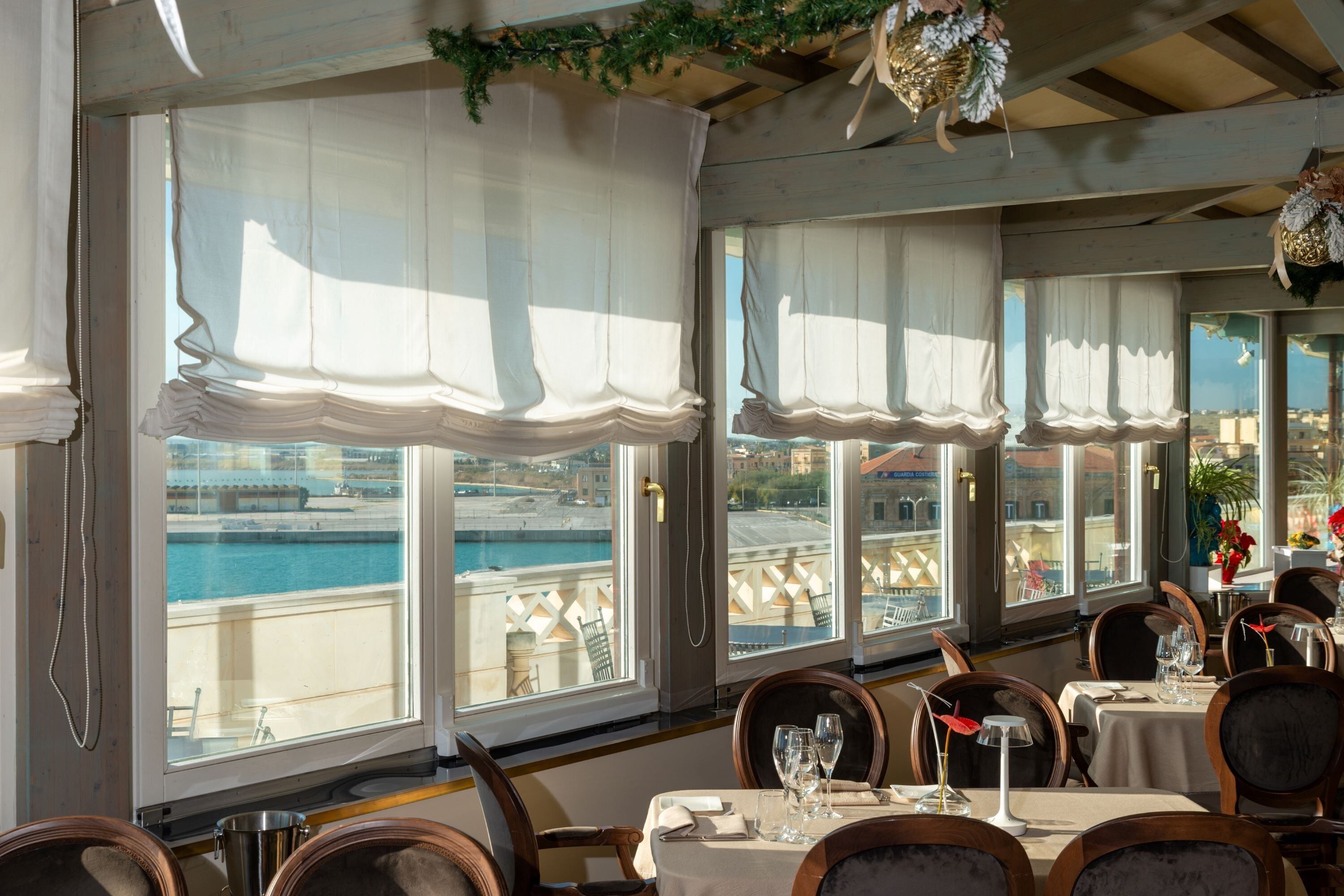 A dining area with tables set for two, large windows with white curtains, and a view of a body of water and buildings.
