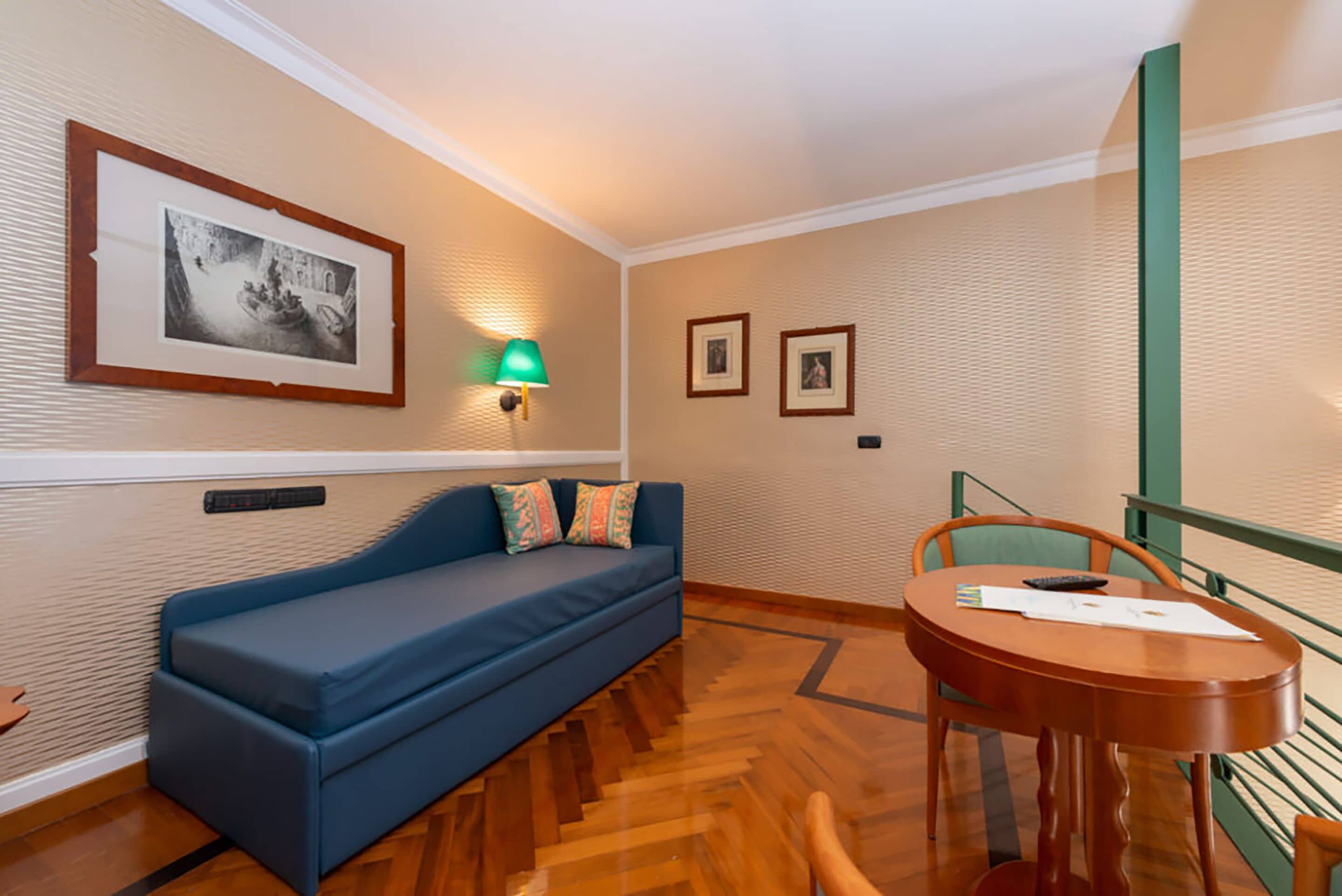A room with a blue sofa, a small table with a chair, a framed picture on the wall, and a lamp.