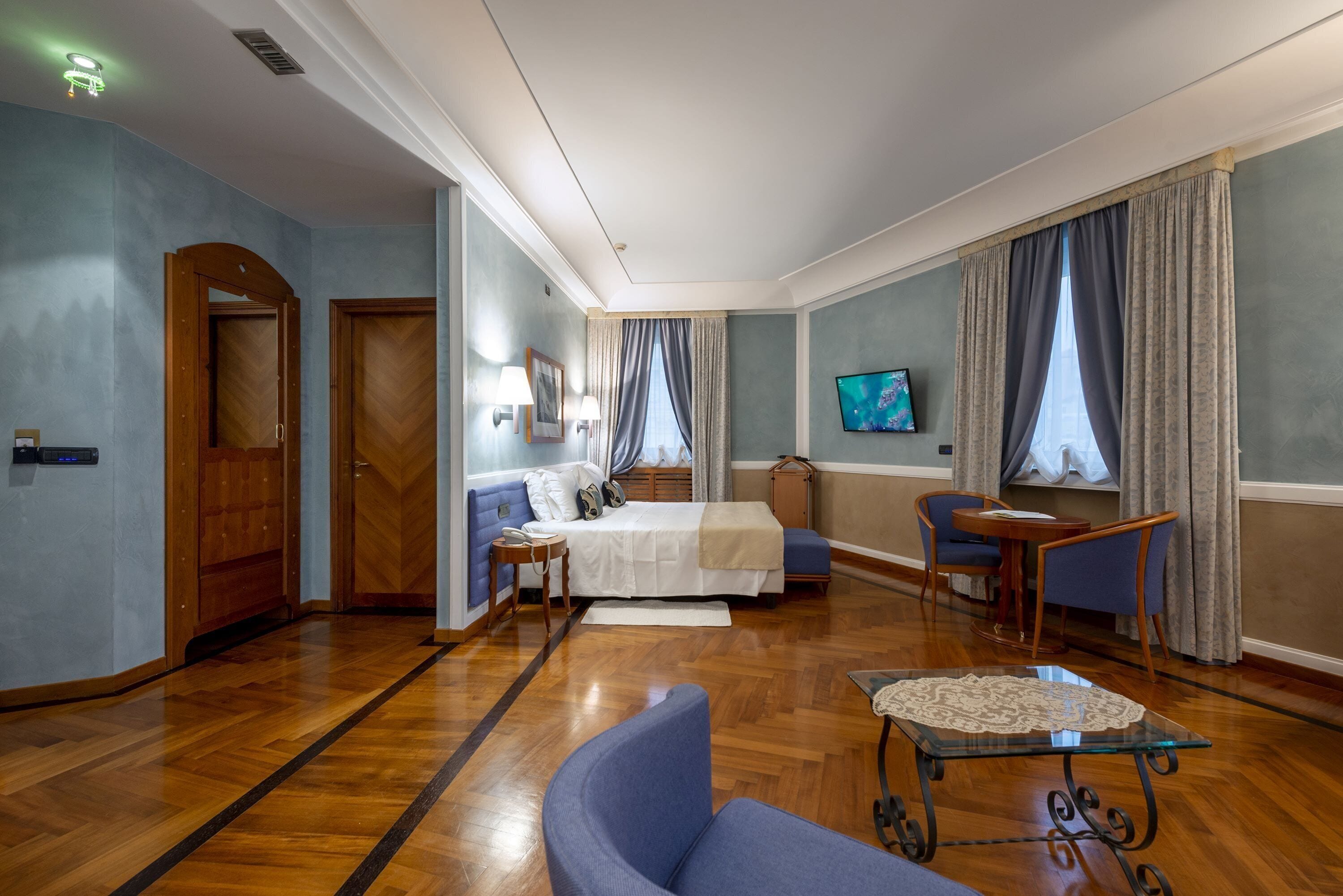 A hotel room with a bed, a seating area with a table and chairs, a TV mounted on the wall, and a wooden floor.